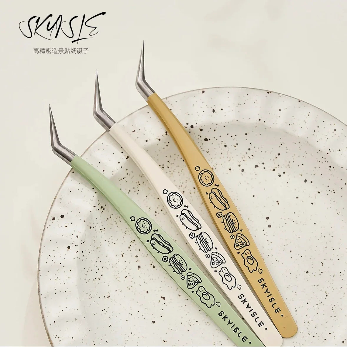 Three angled craft tweezers with food illustrations, resting on a speckled ceramic plate.