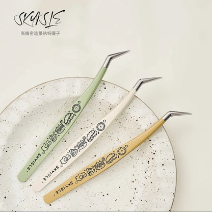 Three pastel angled tweezers with cute food designs for crafting, on a ceramic plate