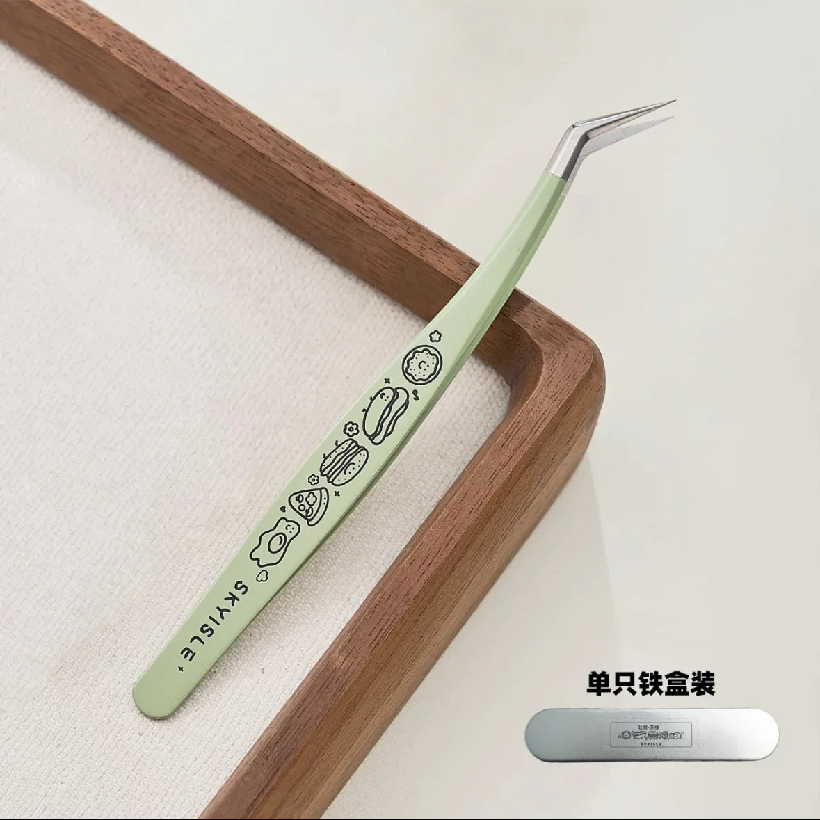 Green precision tweezer with cute food illustrations, angled tip, on white tray.
