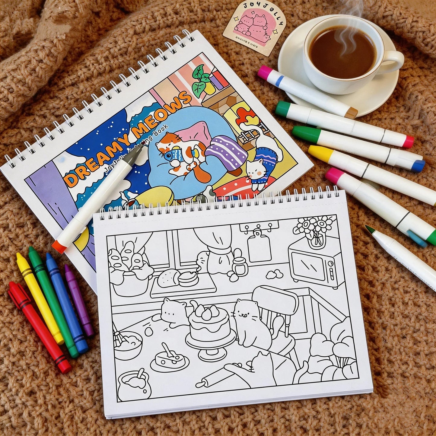 Dreamy Meows cat coloring book with cute cat illustrations, crayons, and markers on a cozy blanket.