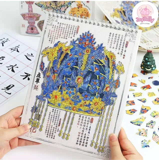 Hand holding a spiral-bound book with intricate blue and gold crown artwork and Chinese calligraphy, surrounded by scene stickers and craft supplies.