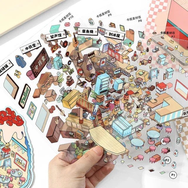 Hand holding clear sheet with colorful city and shop themed scene stickers for journaling or scrapbooking.