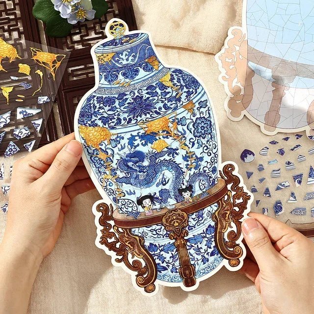 Hands holding a blue and white porcelain vase sticker with dragon design and gold stand, surrounded by decorative sticker pieces for journaling or scrapbooking.