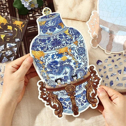 Hands holding a blue and white porcelain vase sticker with dragon design and gold stand, surrounded by decorative sticker pieces for journaling or scrapbooking.