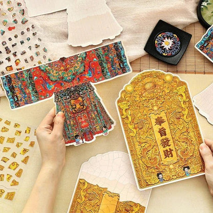 Hands holding vintage-style stickers featuring an ornate imperial silk dance dress and golden dragon-themed plaque with Chinese characters, surrounded by decorative journaling stickers.